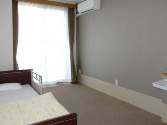エアコンが付いた居室には寝具付きのベッドや洗面所があり、大きな窓にはレースカーテンとカーテンが付いている。