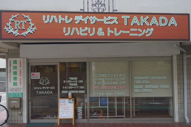 看板のある建物のエントランス リハトレ デイサービス TAKADAの外観 清潔感のあるエントランスです。訪れた方を歓迎する明るく開放的な雰囲気が漂っています。