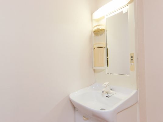 白い壁面沿いに白い洗面台が設置されている。大きな鏡の横に小棚があり、上部に付いている照明が周囲を明るく照らしている。