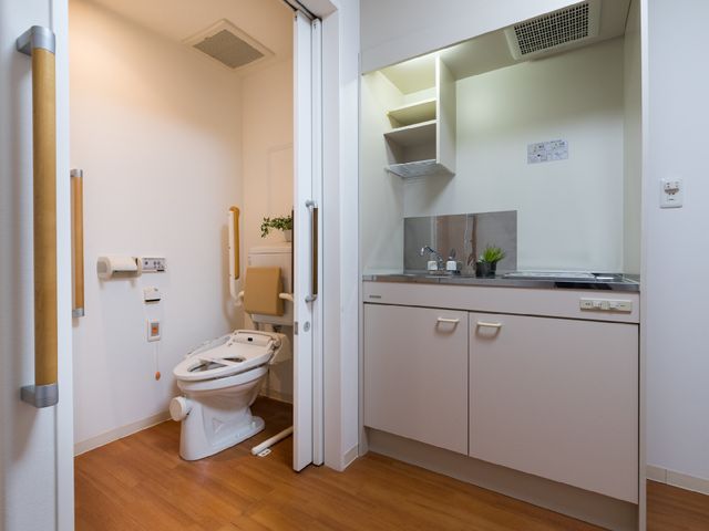 バリアフリーの洋室トイレ 室内 ゆったりとした使い心地のトイレには、安全を考えた手すりが設置されており、清潔感のあふれる空間となっております。小さなキッチンも完備しており、自宅のような安心を提供します。
