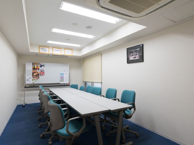 清潔な多目的室 会議室 青いカーペットが印象的な会議室で、壁には掲示物や時計があります。大きなホワイトボードの前には、打ち合わせに最適なテーブルといすが並んでいます。