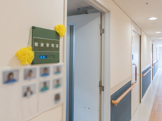 清潔感のある廊下 健康管理室 ゆったりとした廊下は手すりが設置されており、安全に配慮されたつくりになっています。壁には職員の写真が掲載され、アットホームな雰囲気が漂っています。
