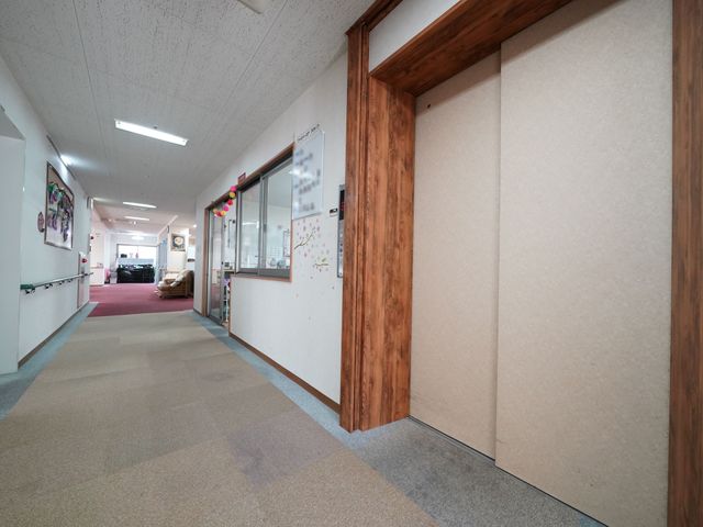 廊下の明るい風景 共有スペース ゆったりとした廊下には木目調の装飾が施され、温かみのある雰囲気を演出しています。そこを行き交う職員の姿が活気に満ちた職場環境を感じさせます。