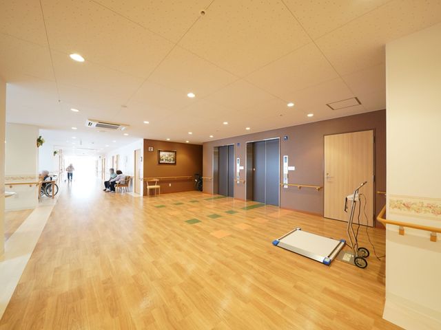 広々とした廊下の一角 共有スペース ゆったりとした通路は木目調の床が心地よく、利用者様がリラックスしながら過ごす様子がうかがえます。車椅子もスムーズに通れる開放的な空間です。