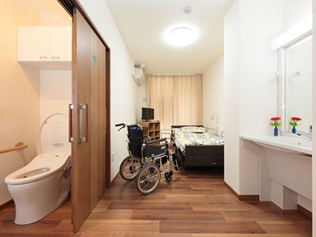 車椅子のある居室 居室   車椅子が置かれた個室で、トイレや洗面台も近くにあり使いやすそうです。木目の床とやわらかな照明で、落ち着いた雰囲気です。
