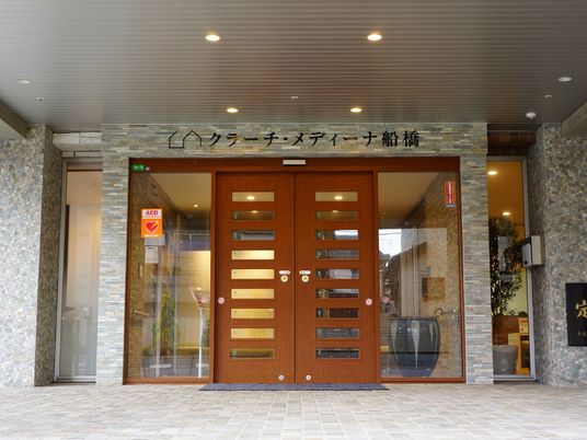 施設の玄関は木とガラスを組み合わせた両開きの自動ドアである。両脇は大きなガラス張りになっており、AEDのシールが貼られている。