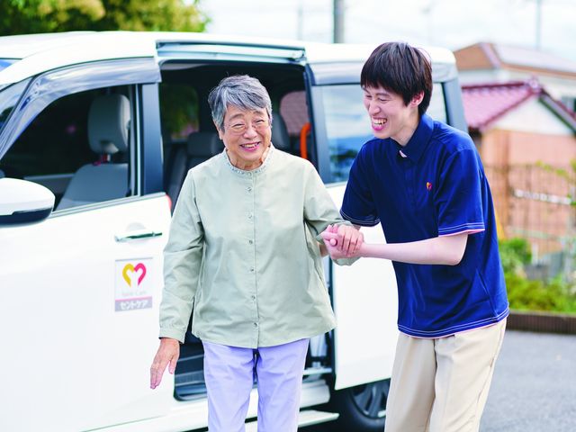 送迎車の利用風景 スタッフイメージ 親しみやすいスタッフが、利用者の方を車まで丁寧にサポートしています。快適な移動をお手伝いします。