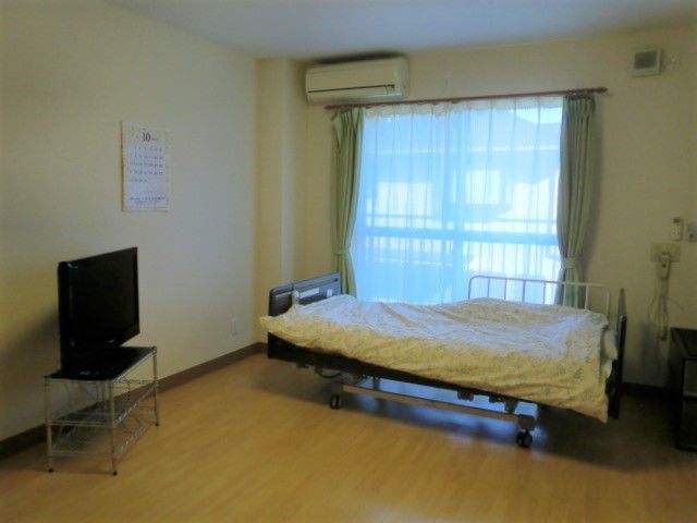 居室のベッドとテレビ シンプルで清潔感のある居室です。窓からの自然光が心地よい空間をつくり出しており、くつろぎの時間を過ごせるでしょう。