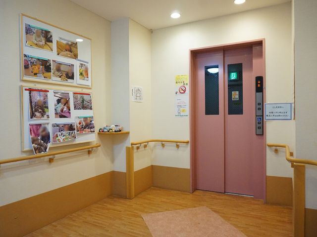 廊下とピンクのエレベーター エレベーター 手すりを設置した暖かみのある廊下には、使い勝手の良いエレベーターが備え付けられています。ボードには日々の活動の様子が写されており、明るい雰囲気が感じられます。