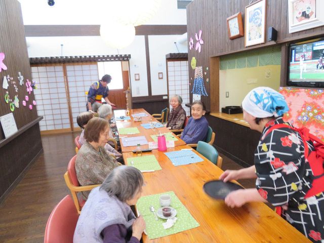 和風ダイニングエリア イベント風景 和風の装いで落ち着いた食堂です。職員が笑顔でお茶を運び、ご利用者はテレビを見ながらくつろいでいます。