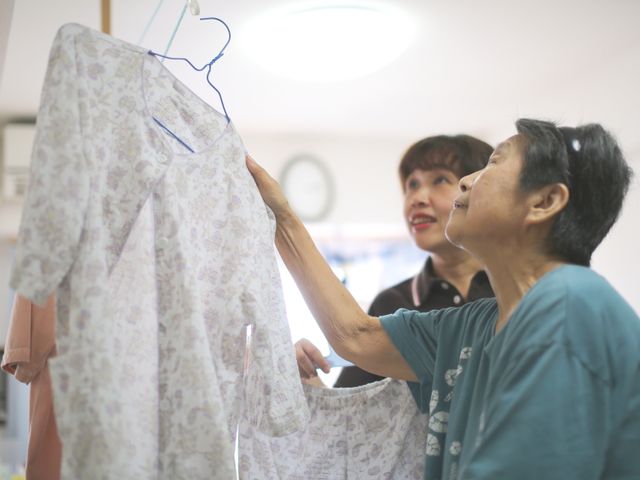 衣類を選ぶ様子の居室 明るい表情で洋服選びを手伝うスタッフを見て、日常生活の支援が丁寧に行われている様子が伺えます。お互いに心地よい時間を共有している姿が見て取れます。