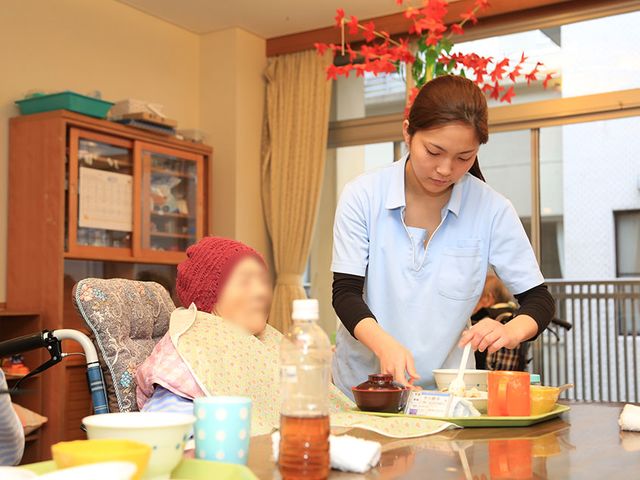 食事を支える様子の食堂 食堂 広い窓から自然光が差し込む明るい空間で、スタッフが入居者に食事を提供しています。