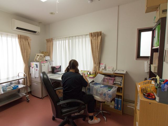 スタッフが業務を行う室内 医務室 整理されたオフィスで、一人のスタッフが書類作業に取り組んでおります。日常の運営を支える、落ち着いた環境です。