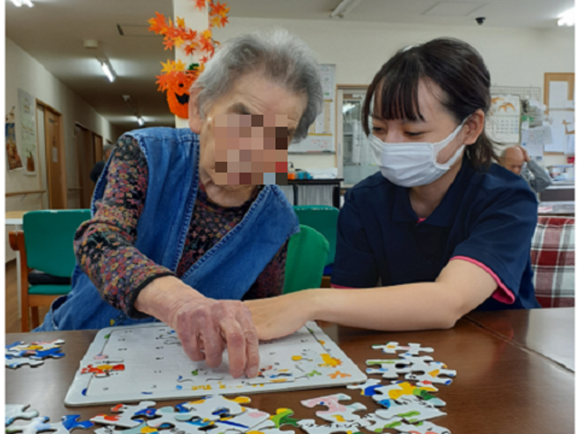 パズルを楽しむ人々の様子 多目的室 笑顔で接するスタッフと一緒にパズルを解く、楽しい活動の一コマです。安心して過ごせる明るい雰囲気が伝わります。
