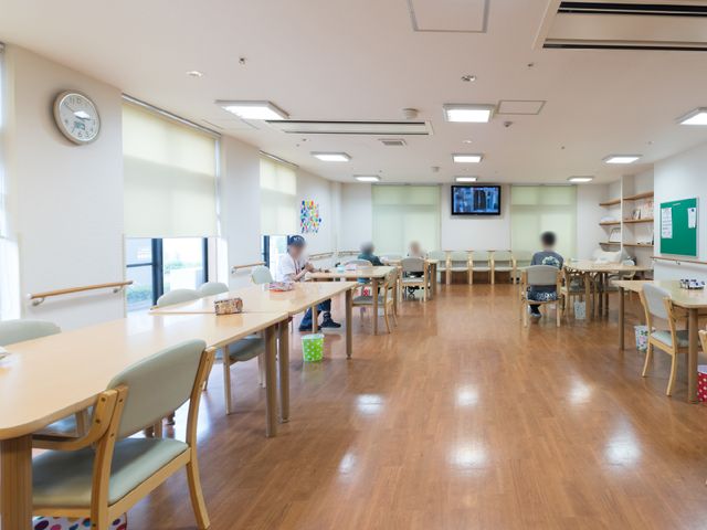 広々とした明るい食堂 ダイニングルーム ゆったりした食堂には、テーブルが整然と配置され、利用者がリラックスしながら食事を楽しんでいる様子がうかがえます。壁には時計が掛けられ、落ち着いた時間が流れています。