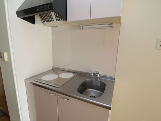 施設の写真 居室内にある小型のキッチンはコンロが２口ある。その上にダクトが設置されている。流し台の上に照明が取りつけられている。