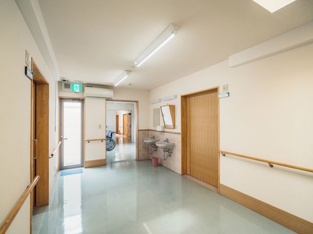 清潔な廊下の一部 通路 広々とした廊下は清潔感があり、手すりが整備されているため歩行に安心です。遠くには車椅子が見え、バリアフリーへの配慮が感じられます。