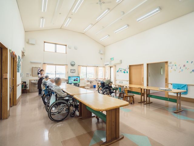 広々とした機能訓練室 食堂 天井が高く、自然光が差し込む機能訓練の場です。スタッフの方が利用者様と丁寧に向き合っている様子が伺えます。