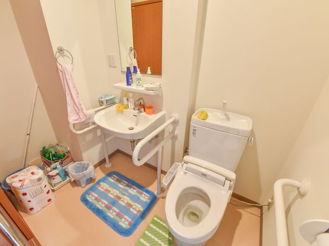 清潔感のある洋式トイレ 施設内トイレ 洋式トイレと洗面台が設置されており、個々のニーズに合わせた快適な環境を提供しています。手すり補助もあり、安心してご利用いただけます。