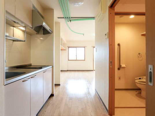 居室内の廊下の左手にキッチン、右手にトイレがあり。奥には居住スペースが広がっている。突き当たりに窓があり、陽射しが入っている。