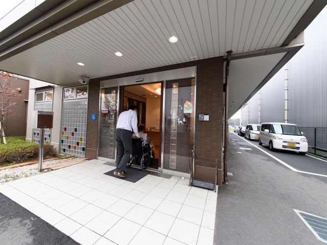 エントランスの状況 玄関 バリアフリー設計の入り口を抜け、スタッフが笑顔で車椅子の方をお迎えしています。働く方にとっても、快適で心地よい環境が整っています。