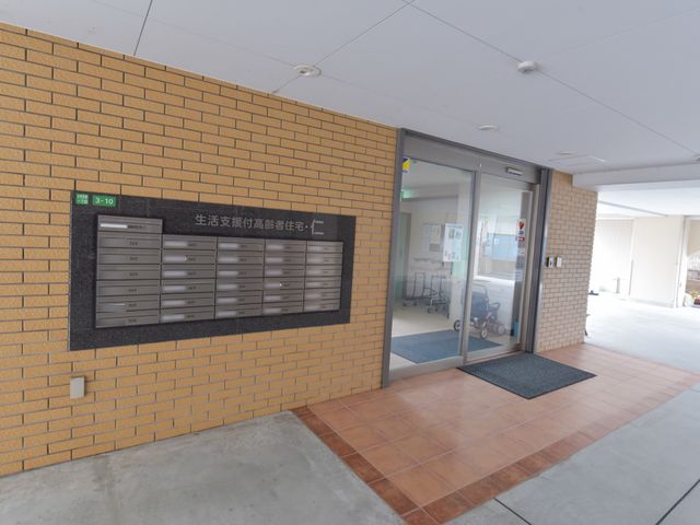 エントランスと郵便ポスト 玄関 ゆったりとした入口には郵便ポストも完備され、明るく開放的な雰囲気を感じられます。遠くに椅子に座る方の姿もあり、心地よい休憩スペースのようです。