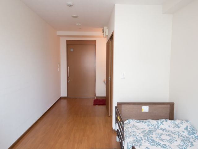 居室のベッドと廊下 居室 ゆったりとした個室で快適な毎日をお過ごしいただけます。手すりの付いた廊下は安心してお歩きいただける設計です。