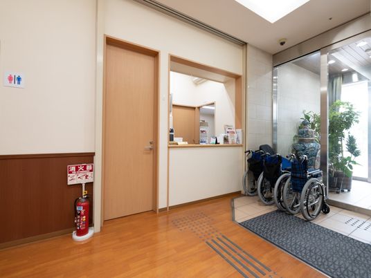 ガラス戸の前にブルーの車椅子が３台置かれている。入口のすぐ横には受付窓口があり、床には点字ブロックが設置されている。