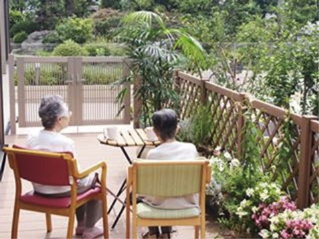 緑豊かなテラス景観 自然風景を楽しむ入居者様 緑に囲まれたテラスで、ご高齢の方が穏やかなひと時を過ごしています。植物と共にリラックスできる、開放感ある空間です。