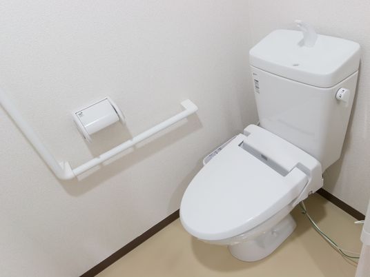 白を基調としたシンプルな個室トイレ。温水洗浄機能付きの便座で、壁にはＬ字型の手すりやペーパーホルダーが設置されている。