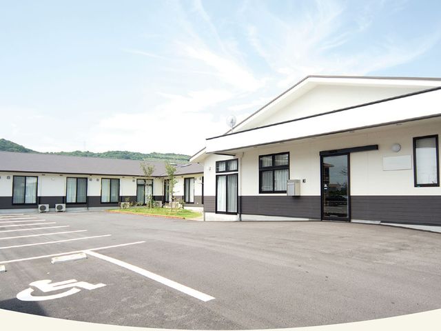明るい建物の外観 外観   ゆったりした駐車スペースがあり、平屋の建物が見えます。青空の下で、落ち着いた雰囲気の外回りです。