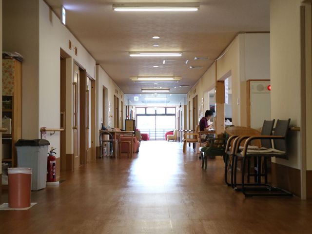 明るい廊下と休憩スペース 廊下 ゆったりとした廊下に沿って落ち着いた色彩の椅子が配置され、一人掛けのスペースでスタッフが書類に目を通しています。