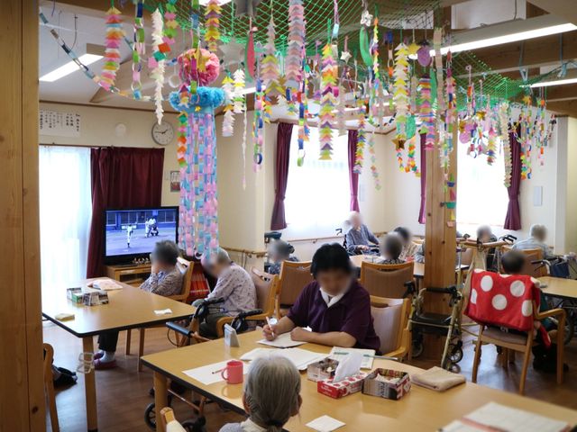 装飾の多い居室空間 共有スペース 色とりどりの飾りが天井から下がる居心地の良い空間です。テレビを見ながらくつろいでいる方々の姿が印象的です。