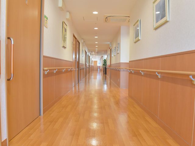 明るく清潔な廊下 施設内廊下 床は滑りにくい素材で、壁には手すりがついており、遠くに一人の方が歩かれています。安心して歩ける環境が整っていますね。