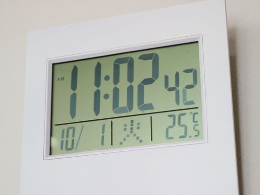 白いフレームのデジタル時計が白い壁に掛けられている。時刻、日付け、曜日、室温がデジタル表記されている。