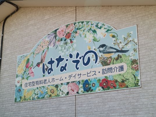 施設建物の壁面に施設プレートが設置されている。花や鳥が描かれたプレートに施設名称と併設するサービス事業名が記載されている。