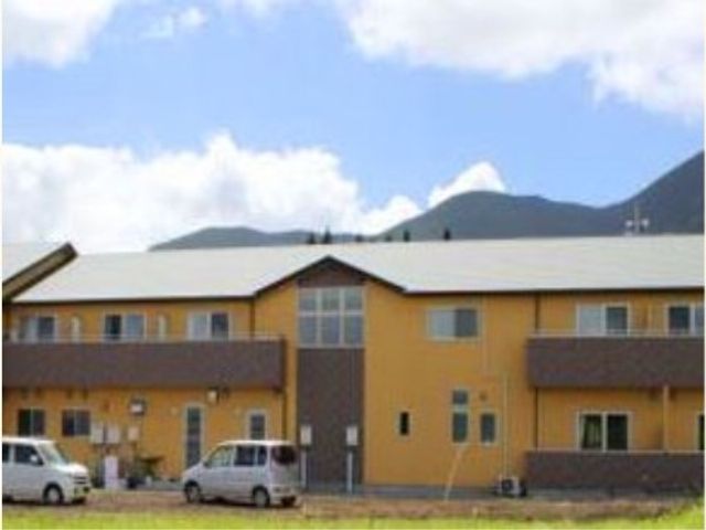 山並み背景の黄色い建物 外観 広々とした駐車場を有する、のどかな山々を背景にした明るい色合いの建物です。自然光あふれる環境で、快適な勤務が可能です。