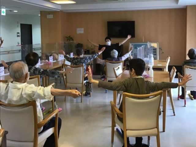 運動中のリビングの様子 体操 ゆったりとしたリビングで、利用者の皆様が健康体操を行っており、笑顔でエクササイズに取り組んでいます。安全で明るい空間で、体を動かすことの楽しさを共有できる場所です。