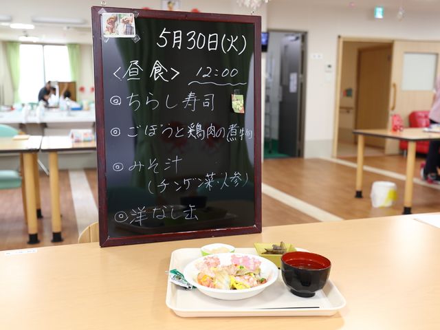 明るい食堂の様子 献立を書いたボード 手書きのメニューが暖かい雰囲気を醸し出し、背景にはスタッフが見守る清潔感のある食堂が広がっています。