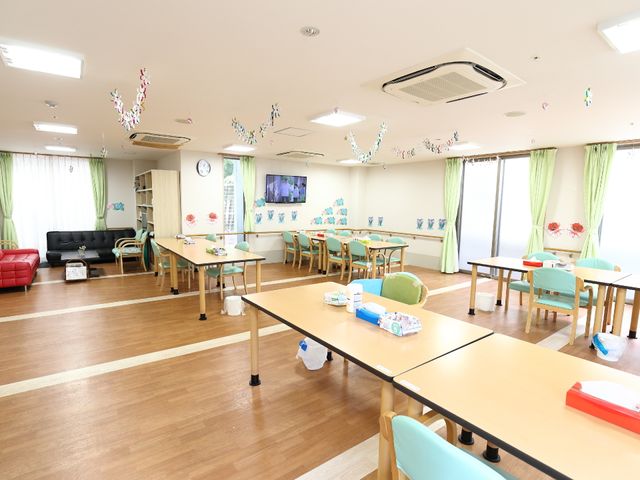 広々とした共有リビング リビングルーム 広々としたラウンジは窓から光が差し込み、様々なアクティビティが楽しめる明るい空間です。ゆったりとしたソファやテーブルが配置されており、利用者様がくつろぐ姿が想像できます。