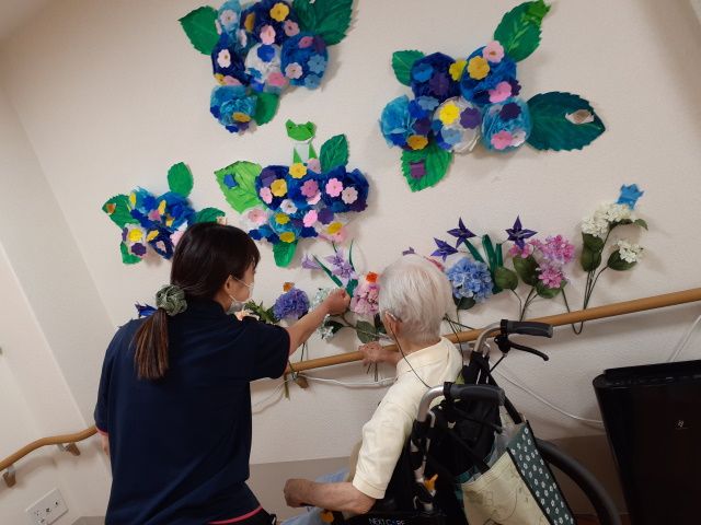 花壁画で楽しむ居室 居室 壁に飾られた花の壁画を前に、利用者とスタッフが楽しげに会話をしています。この明るく温かな雰囲気は、ご家族にとっても安心のポイントです。