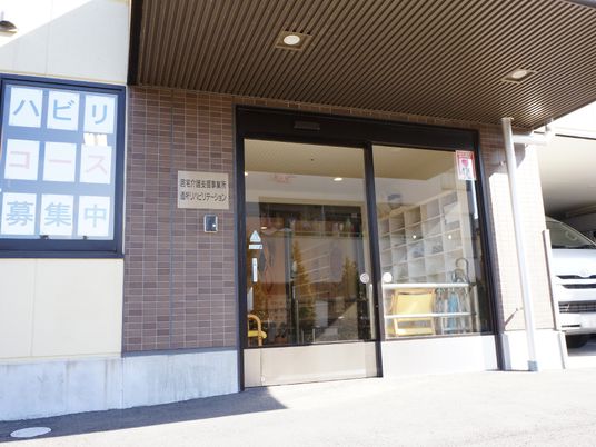 施設の入口部分には、大きめの自動ドアが設置されている。雨除けの屋根があり、「リハビリテーション」という掲示物が目立つ形で掲げられている。