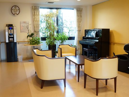 テーブルの周りには3つのイスを確認することができる。また周りには大小の観葉植物や、ピアノ、マッサージ機などもある。