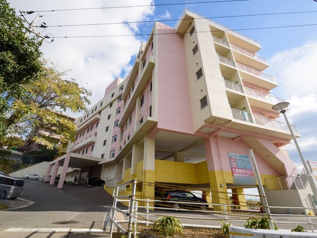 ピンク色の多階建て外観 外観 高い立地にある明るいピンク色の建物です。広々とした駐車スペースが確保されており、利便性に優れています。