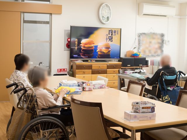 共有スペースの日常風景 共有スペース 明るいリビングで居住者同士がくつろいでいます。テーブルを囲みながら、テレビを視聴したり、お喋りを楽しんだりする様子が伺えます。