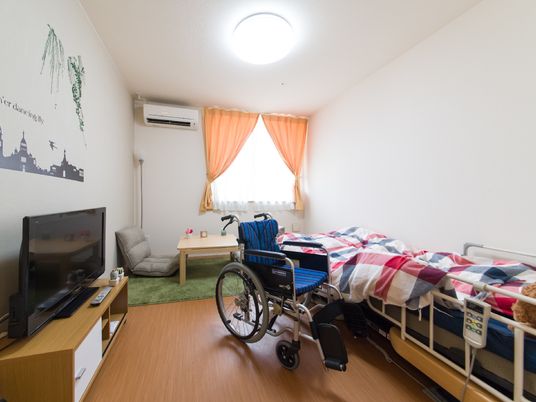 白い壁に車椅子のままでも利用しやすいフローリングの縦長の部屋。自然光の入る窓がある。エアコン、介護用ベッドも完備している。