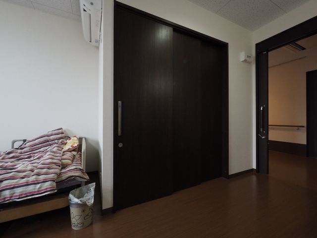 居室のベッドと扉 トイレ扉 シンプルで清潔感のある居室は、個人のプライバシーが確保されており、心地よい生活空間を提供しています。ベッド周りは十分なスペースがあり、動きやすい造りになっています。