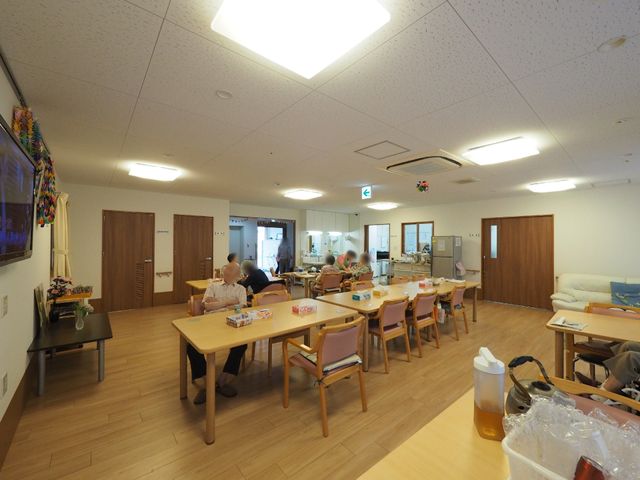 共用スペースの様子 食堂 広々とした空間で利用者がくつろいでおり、一角には大型テレビも設置されております。椅子に座ったり、テーブルを使用して様々な活動が行える環境が整っています。