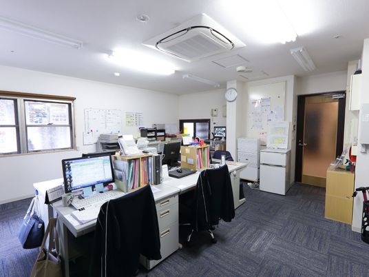 白色の天井と壁にデザイン仕様の床の内装である。中央に寄せてデスクが並べてある。その上にはパソコンやファイルなどが置かれている。
