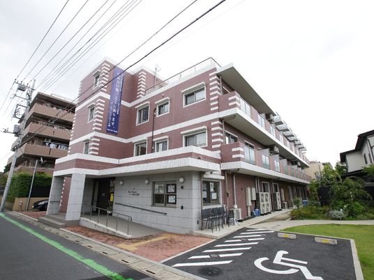 道路に面したグレーと茶系のタイル調の外壁の3階建て建物である。建物の傍には車椅子用の駐車スペースがある。